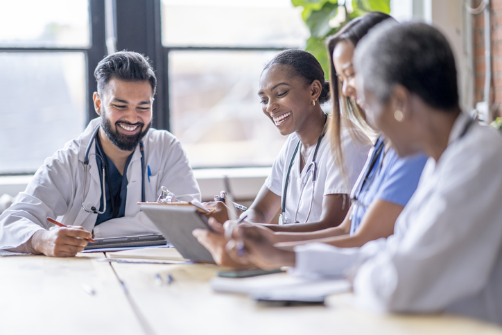 A small group of four medical professionals sit around a boardroom table as they meet to discuss patient cases. They are each dressed professionally in scrubs and lab coats as they focus on working together.